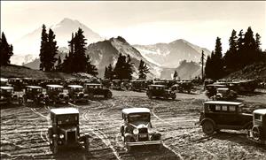 Many automobiles from the early twentieth century are parked in a dirt clearing. Tall evergreens and a large snowy peak are in the background. 